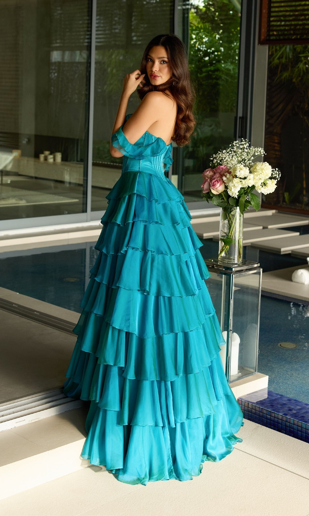 A woman in the turquoise Off-Shoulder Ruffled Prom Gown Ava Presley 42213 stands by an indoor pool, looking over her shoulder with a vase of flowers on a glass table beside her.